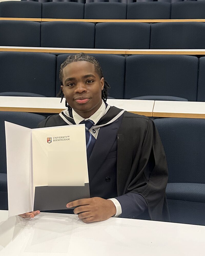 Daniel at his graduation, in an academic gown over a suit and tie, holding an open degree certificate while seated in a lecture theatre